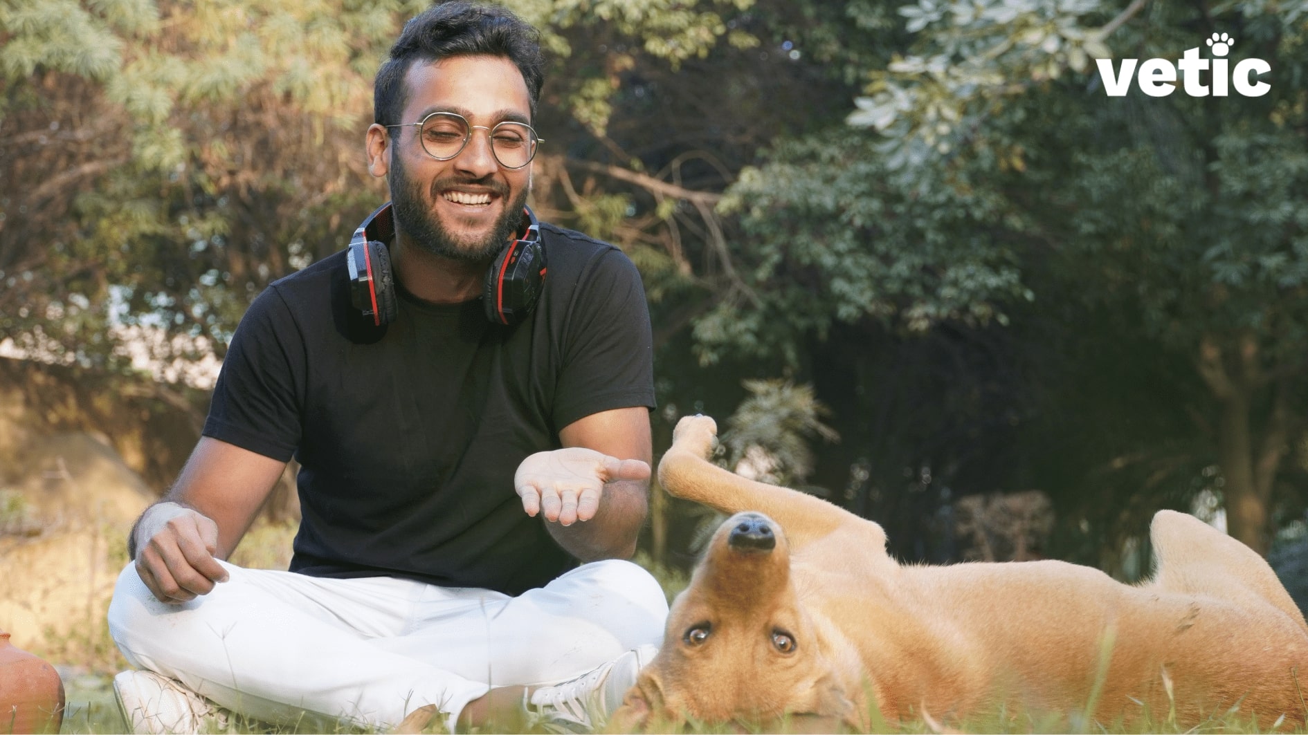 Happy and smiling man in black t-shirt with headphones around his neck sitting on the grass playing with his dog who's laying on the grass.