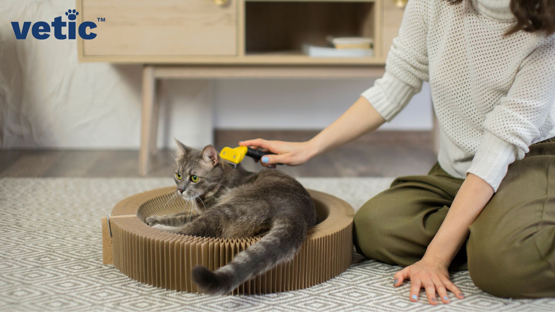 Closeup of a brown-grey ticked cat on a cat scratcher being brushed by their owner with a yellow deShedding brush