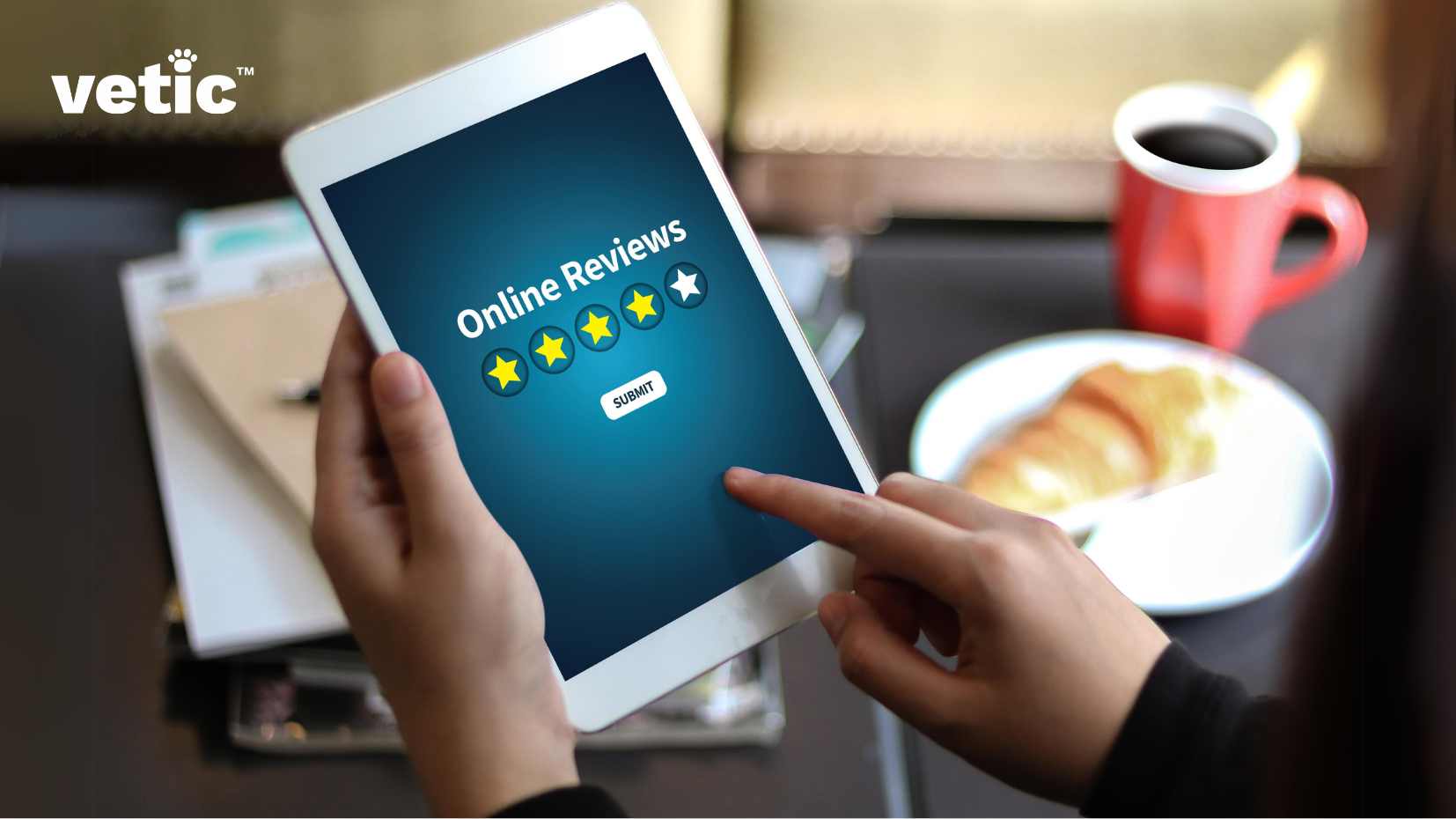 The image depicts a person holding a tablet displaying an online review page with a five-star rating system. A red coffee mug and a plate with a croissant are also visible in the background. The central focus is on a person holding a white tablet, which displays an “Online Reviews” page. You should check the online review of each groomer to find the best dog groomer near you. On the tablet screen, there are five yellow stars indicating the rating system and a “submit” button. The person’s hand is visible, pointing at the tablet screen. In the background, there is a red coffee mug filled with coffee placed on dark surface. Next to the coffee mug, there’s also a white plate with what appears to be a croissant on it. There are some papers and folders under the tablet suggesting an office or work environment.