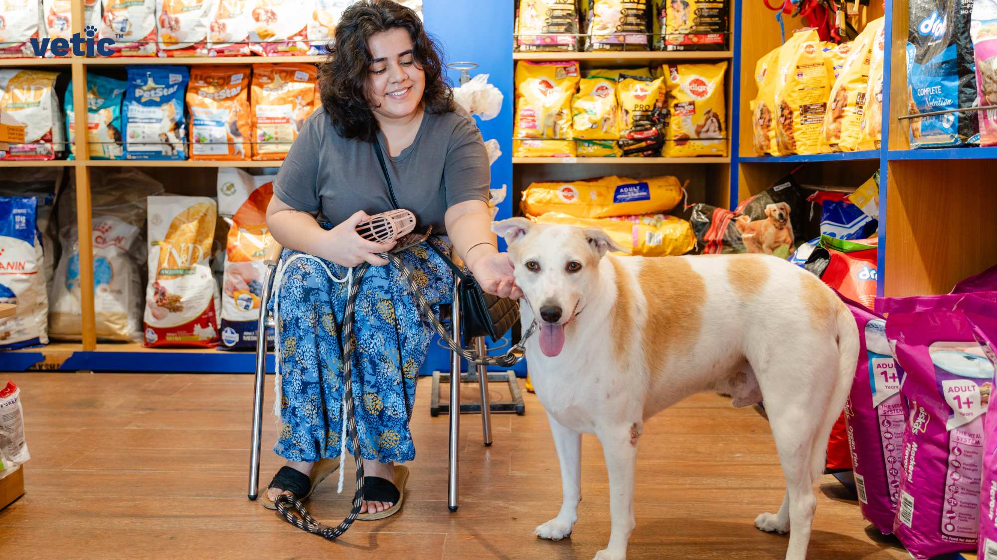 Certainly! The image depicts an individual sitting on a bench inside a store with pet supplies. The individual is holding a leash attached to a large, light-colored dog standing next to them. The dog appears to be looking at the camera with its mouth open, possibly panting. Behind them are shelves stocked with various bags of pet food and other pet-related products. The image by Vetic is a perfect depiction of how to handle your pet while visiting the vet